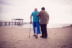 Man assisting elderly man with walking on the beach, an example of patient-centered care.