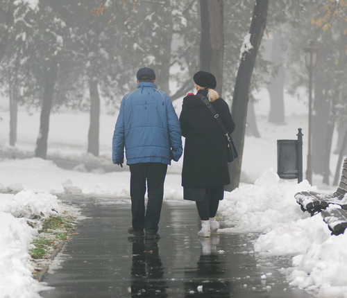 Two elderly people walking on the sidewalk during winter with snow on the ground.