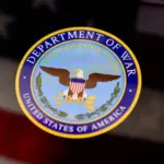 The United States Department of War seal is displayed on a smartphone screen. In the background^ an out-of-focus American flag is visible. This conceptual image illustrates themes of US military history^ national defense^ government institutions^ and historical conflicts.