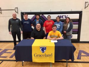 West Central Valley senior Alex Kalbach (right yellow t-shirt) puts pen to paper and signs a letter of intent with Graceland University on February 19. Photo courtesy of Dave Doely/Alex Kalbach.