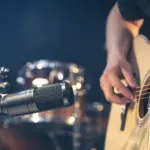 Male musician playing acoustic guitar behind microphone in recording studio.