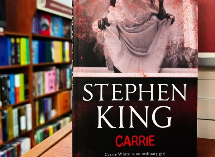 Paperback cover of Carrie by Stephen King shown on a bookshop shelf background