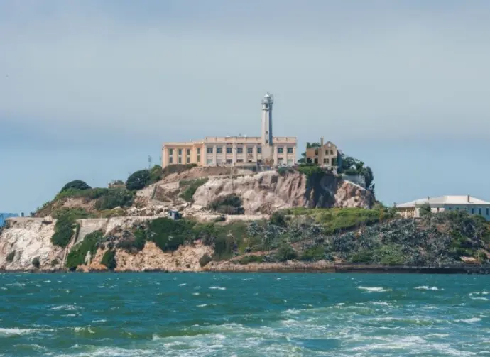 Alcatraz Island in San Francisco Bay^ California^ USA^ viewed from the water. Shows rugged terrain^ prison building^ lighthouse^ and choppy water under clear sky.