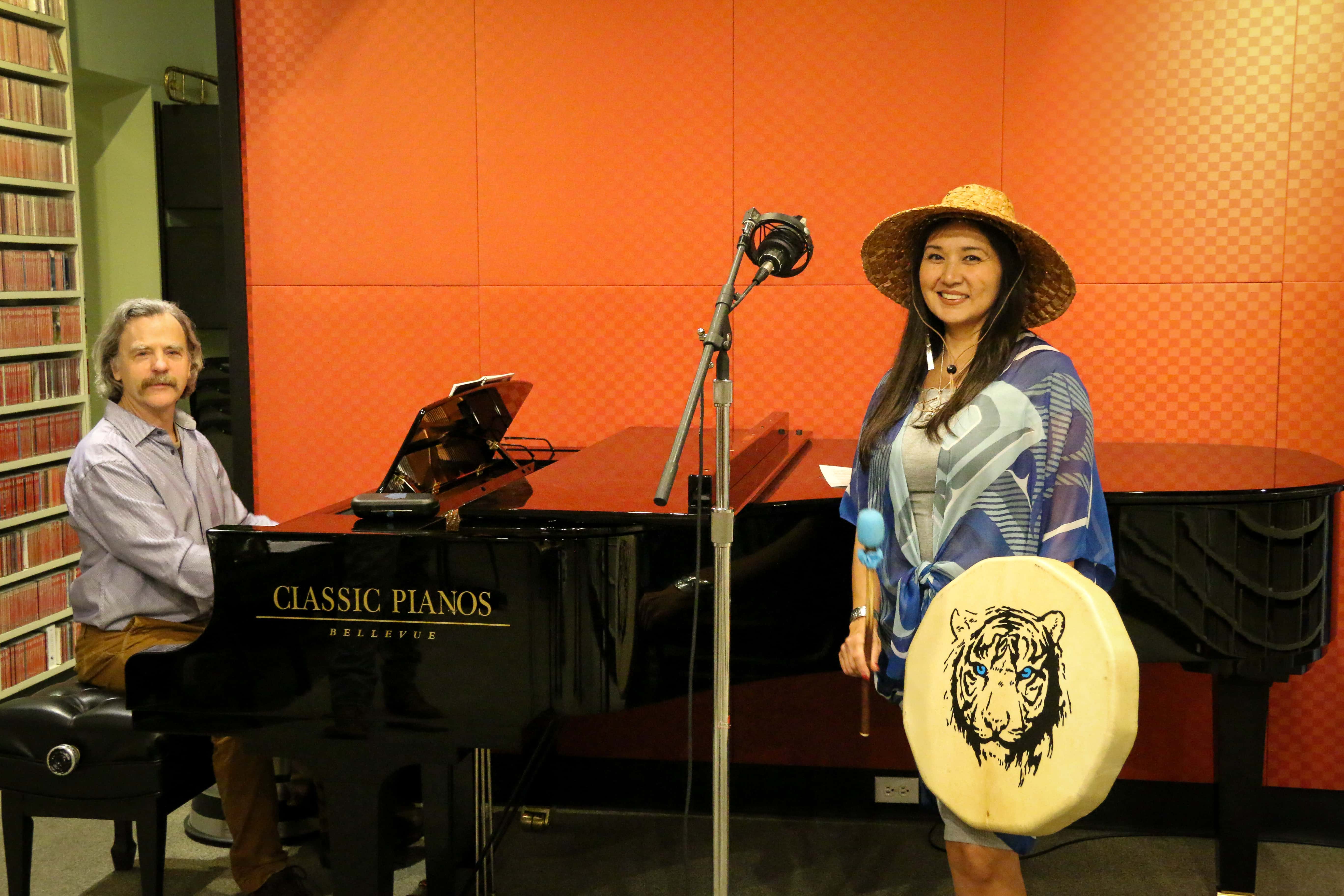Woman in Alaska Native attire stands near a microphone, holding a drum, with a man seated next to her at a grand piano