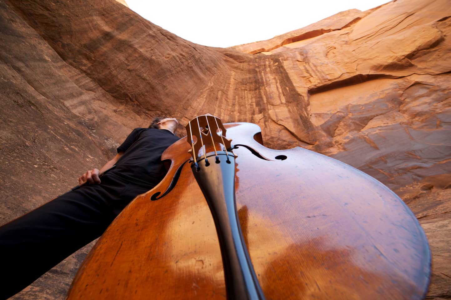 Close-up view of upright bass with man standing behind, and tall rock formation beyond (photo by Gabriel Gomez)