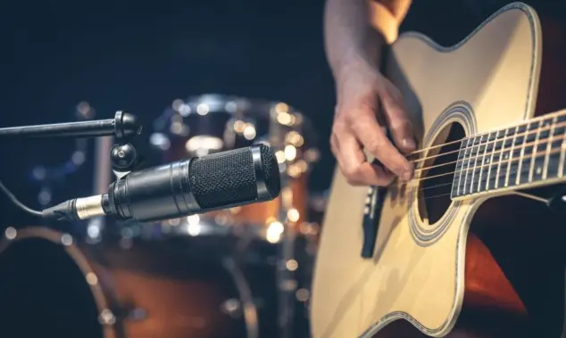 Male musician playing acoustic guitar behind microphone in recording studio.