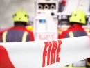 Scene of incident is cordoned off with red and white tape^ reading ‘fire’. Out of focus in the background are three firefighters^ two fire engine appliances and smoke billowing through the air.