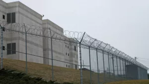 Looking up at the outside walls of a modern prison surrounded by a fence and barbed wire.