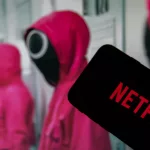 Netflix logo on the display of a smartphone in front of a television with the new series "Squid Game" ( focus on Netflix logo )