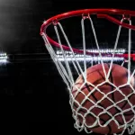 Looking up at an orange basketball falling through the rim and a white nylon net. With the arena lights in the background.