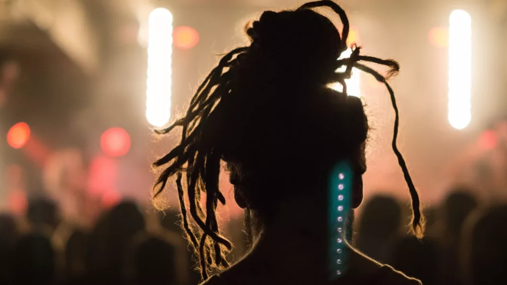 Close up portrait of an anonymous performer/ silhouette with dreadlocks, back to camera, watching a concert on stage with music, retro light effect, flashlight and spotlight in a festival.