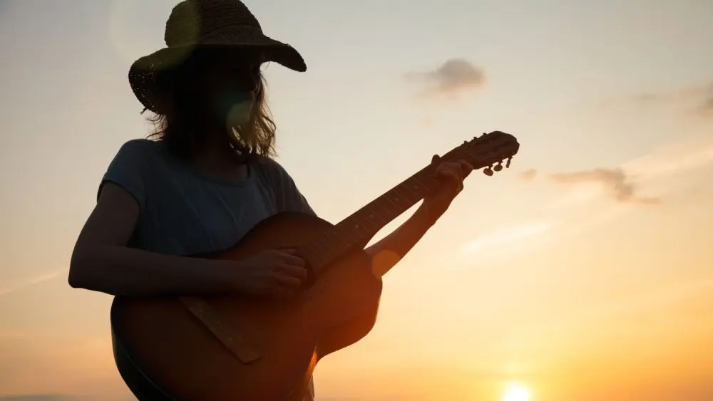 silhouette of young free woman in straw hat playing country music on a guitar at sunset, copy space