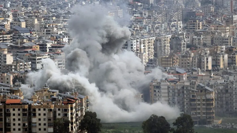 Large plumes of grey smoke billow from mult story apartment buildings following an explosion in a densely populated city, depicting the devastation of modern urban warfare. Jan 28 2026 in USA