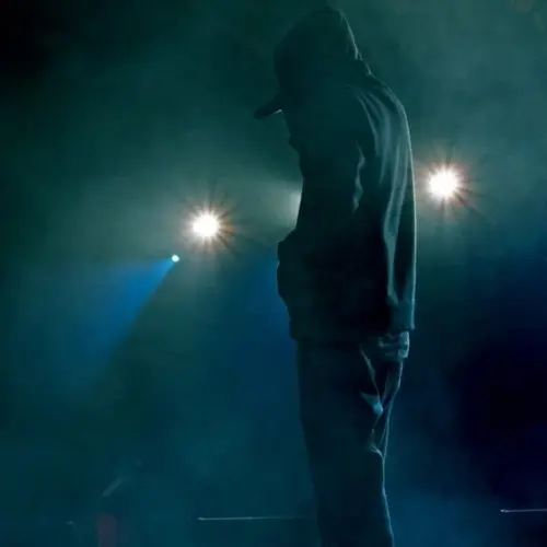 Rap artist performing on stage under soffit lights. Concert backlighting and illumination during live music show. Singer in hoodie holding microphone^ starting the concert