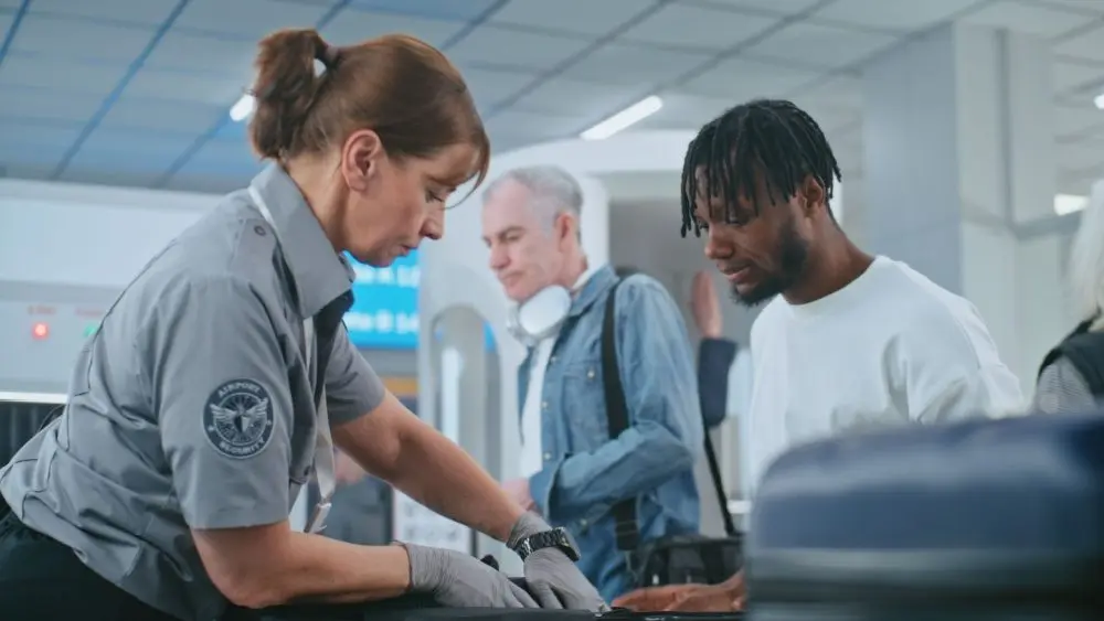 Security Checkpoint in Airport Terminal: Female TSA Worker Inspecting Baggage of Passenger before Boarding Flight^ Finding and Confiscates Liquid. Queue of Diverse People During Screening Procedures.