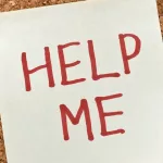 sticky note with 'help me' written in text in front of a cork board background