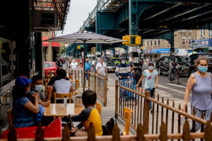 Pedestrians wearing protective masks pass in front of customers sitting outside to eat at a restaurant in the Corona neighborhood in the Queens borough of New York, U.S., on Saturday, June 27, 2020. New York's public transport usage has risen to almost half the typical level before the coronavirus outbreak, a recovery that has yet to extend to the restaurant industry.