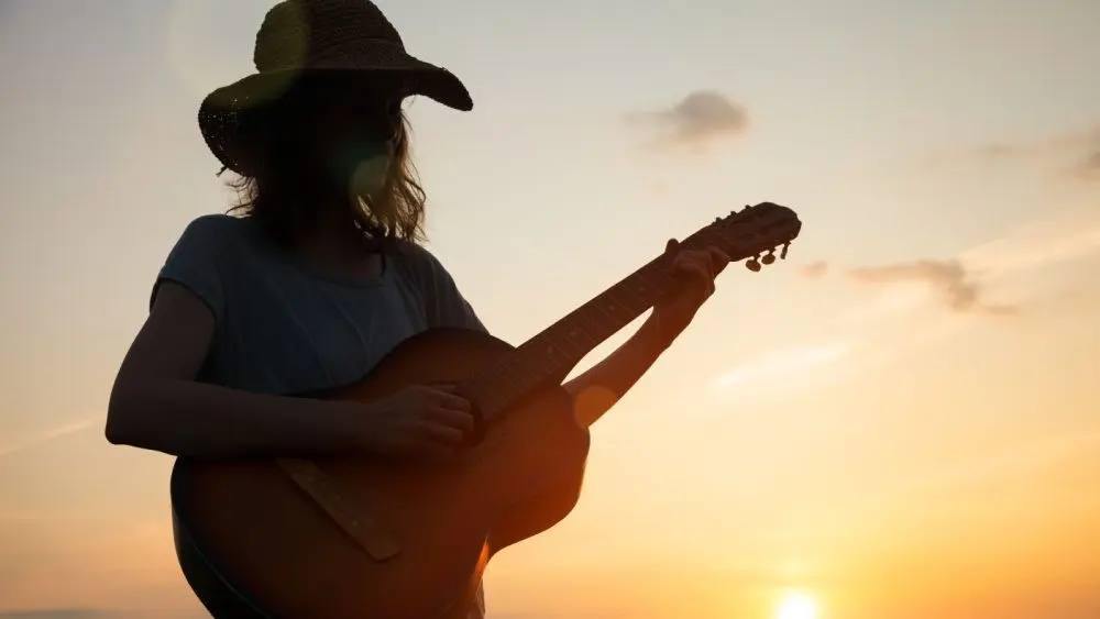 silhouette of young free woman in straw hat playing country music on a guitar at sunset^ copy space