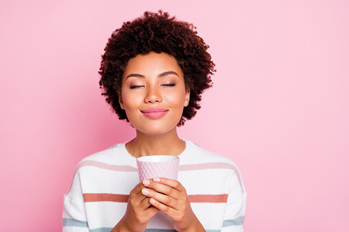 Photo of pretty dark skin lady holding in hands hot beverage cup dreamy closing eyes enjoy good smell wear striped pullover isolated pastel pink color background