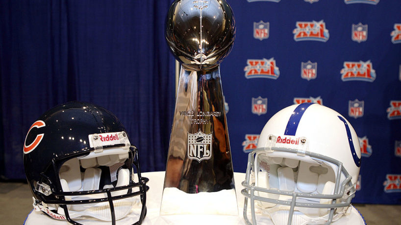 The Vince Lombardi Trophy sits between the Chicago Bears helmet on the left and the Indianapolis Colts helmet on the right on a table in front of an NFL backdrop