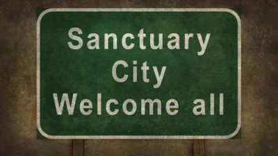 Green highway sign saying "Sanctaury City Welcome All).