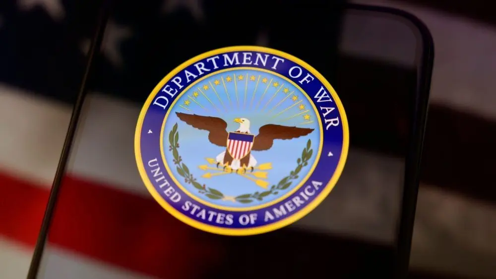The United States Department of War seal is displayed on a smartphone screen. In the background^ an out-of-focus American flag is visible. This conceptual image illustrates themes of US military history^ national defense^ government institutions^ and historical conflicts.