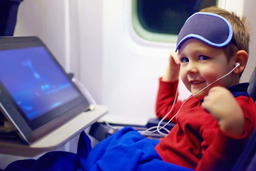 Kid with an ipad and eye covers in the middle seat of an airplane,