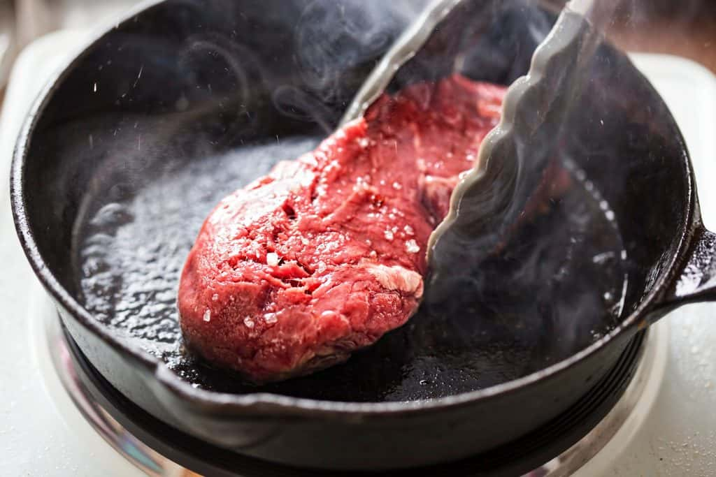 An image of steak being seared in a pan with tongs flipping it