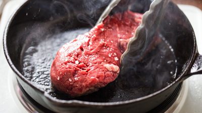 An image of steak being seared in a pan with tongs flipping it
