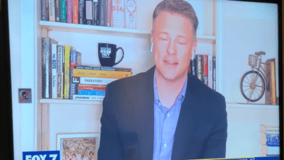 Fox 7 news anchor Dave Froehlich broadcasting from home in front of unleveled shelves