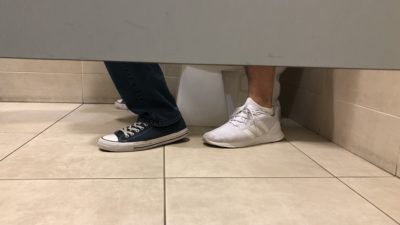 view of a toilet stall showing two pair of feet implying two people are on the toilet at the same time