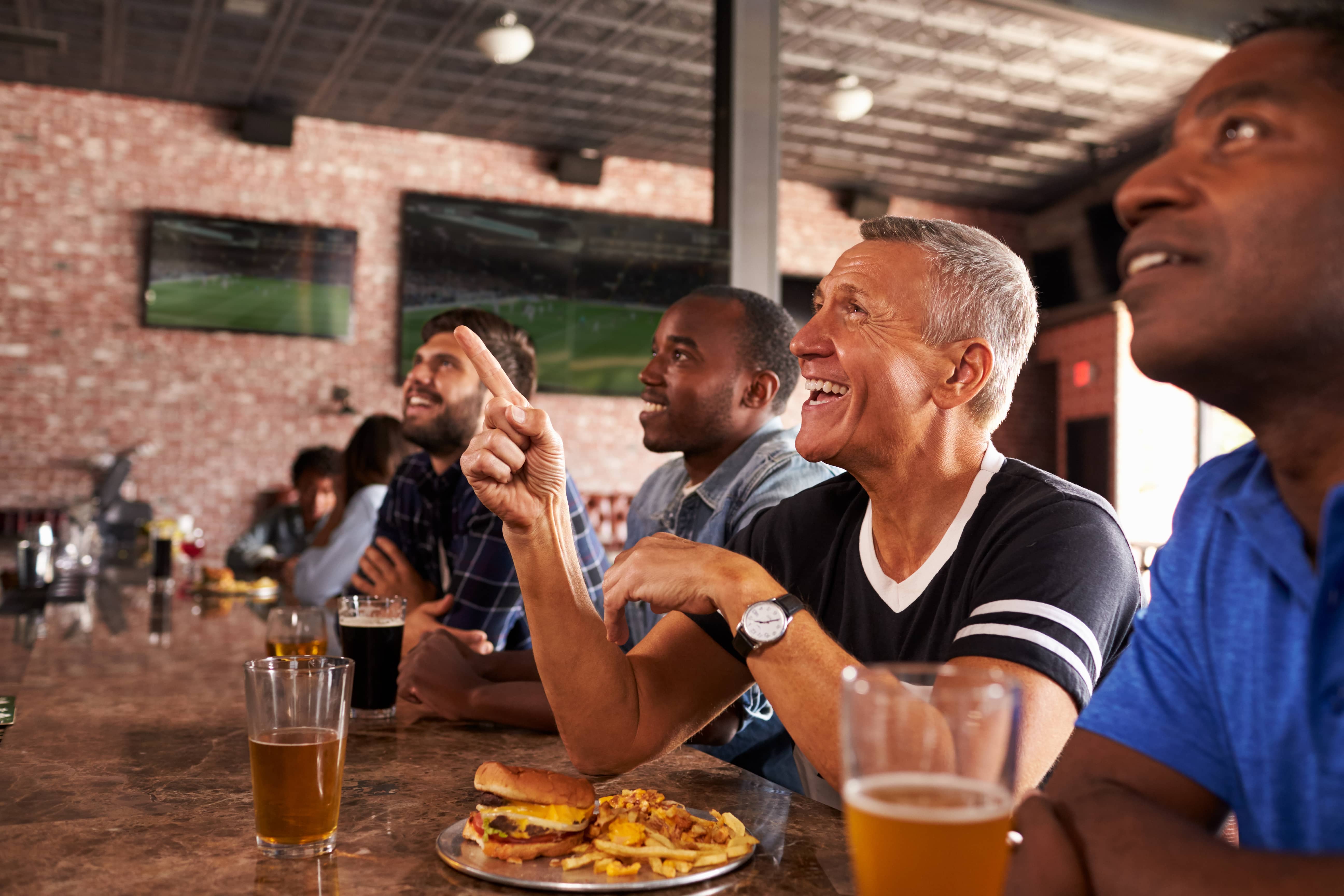 Male Friends At Counter In Sports Bar Watching Game