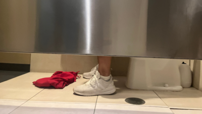 Nick's feet seen from under the bathroom stall, with a pair of shorts lying in front of him.