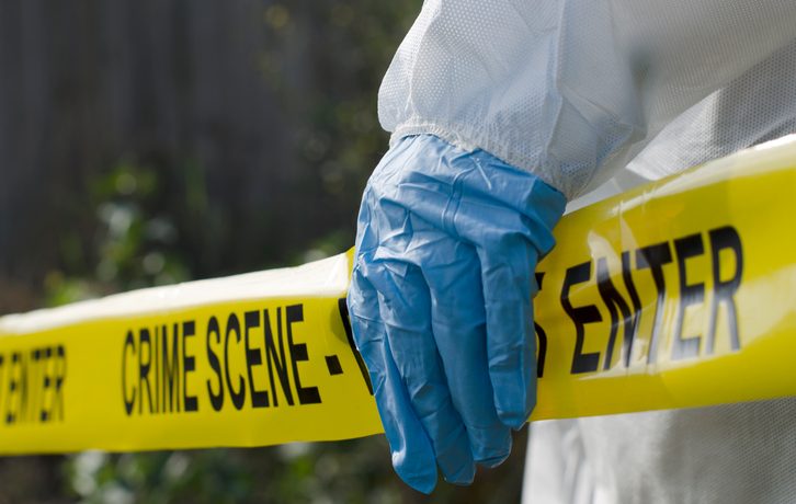 A blue-gloved hand grips a piece of yellow crime scene tape. Photo by Paul Fleet.
