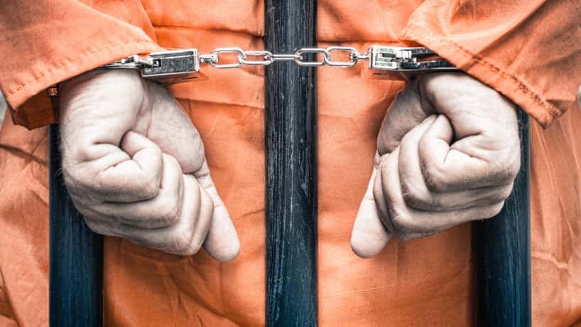 A handcuffed person in an orange jumpsuit resting against prison bars.