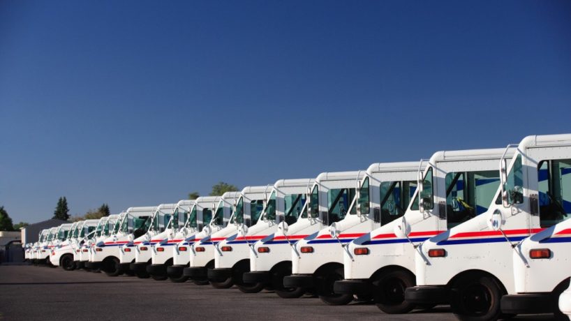 A fleet of U.S. postal trucks sits in a line outside of the USPS center.