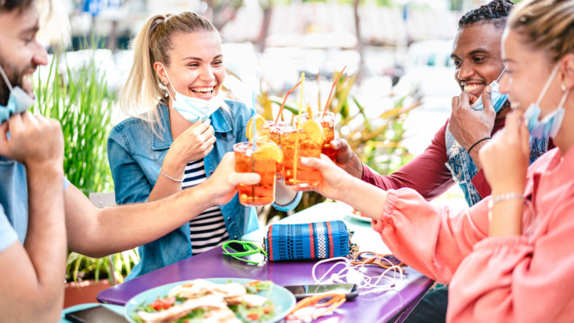 Friends drinking spritz at cocktail bar with face masks - New normal friendship concept with happy people having fun together toasting drinks at restaurant - Bright filter with focus on left woman