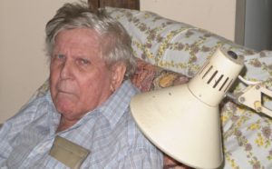 Grumpy Senior Man sitting on a comfy chair with a cushion behind him and an old lamp beside him, in Queensland, Australia