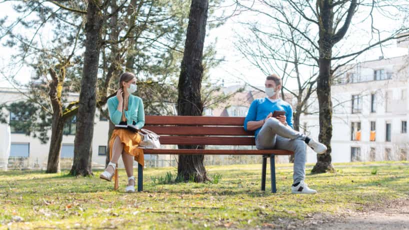 Woman and man with face mask in social distancing sitting on a park bench