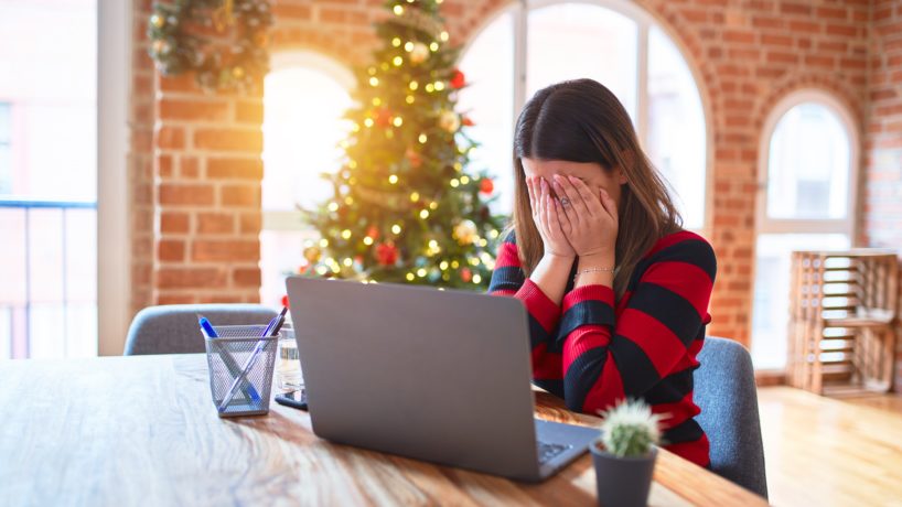 woman sitting at the table working with laptop at home around christmas tree with sad expression covering face with hands
