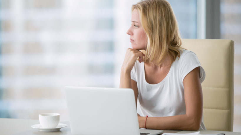 Young attractive woman at a modern office desk, working with laptop, looking at the window, thinking about a post, full-time blogger, seeking for inspiration, help to be productive, updating computer