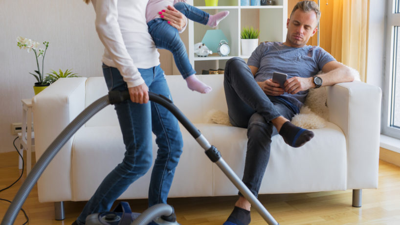 Woman with small child in her hands doing housekeeping while man sitting in couch and relaxing.