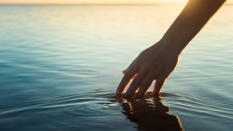 A female hand touching the ocean water in front of a beautful sunset during summer tim