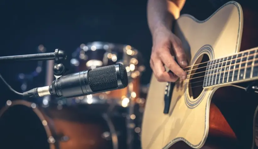 Male musician playing acoustic guitar behind microphone in recording studio.