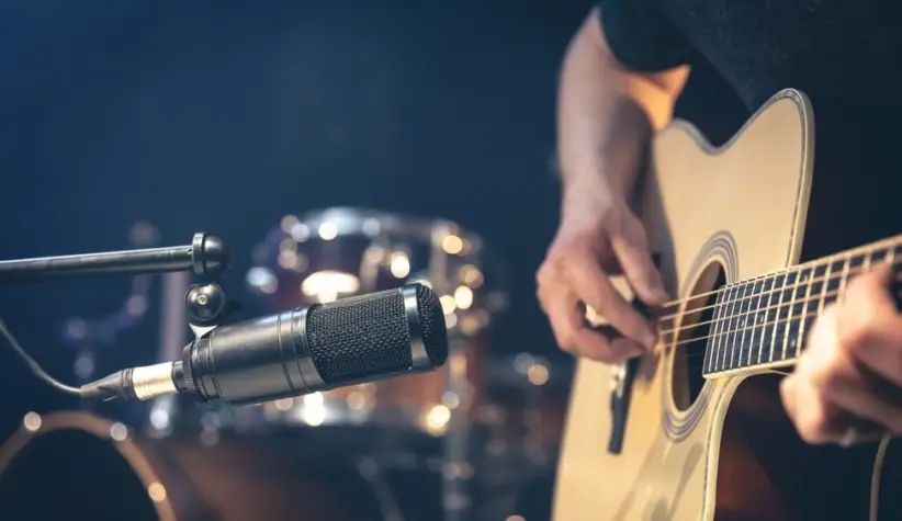 Male musician playing acoustic guitar behind microphone in recording studio.