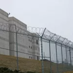 Looking up at the outside walls of a modern prison surrounded by a fence and barbed wire.