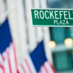 Close-up view of green street sign depicting it is Rockefeller Plaza in Midtown Manhattan^ New-York. Blurred American flags in the background