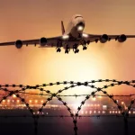 Passenger plane takes off at dusk^ in the background the lights of the airport can be seen and in the foreground a barbed wire fence