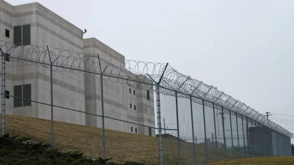 Looking up at the outside walls of a modern prison surrounded by a fence and barbed wire.