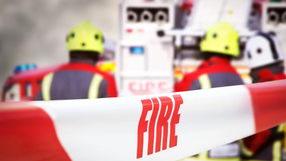 Scene of incident is cordoned off with red and white tape^ reading ‘fire’. Out of focus in the background are three firefighters^ two fire engine appliances and smoke billowing through the air.